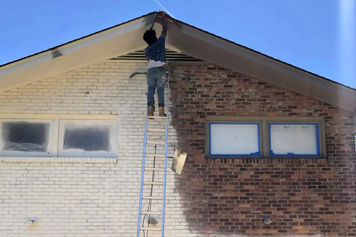 Our painter on a ladder spraying the exterior of a brick home, with windows and garage doors masked off