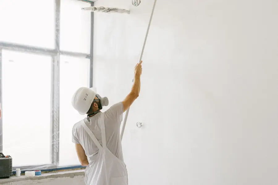 Painter in white work clothes and a mask rolling a wall with an extension pole