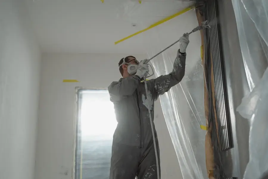 Painter in coveralls and a respirator spraying an interior wall, with the doorway masked off in plastic